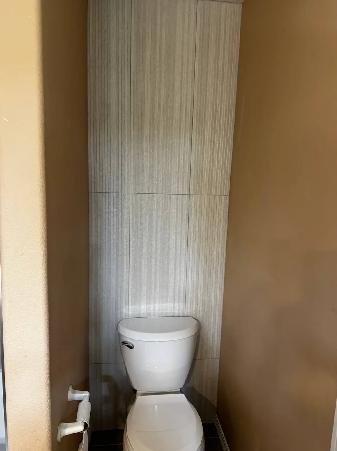 Floor-to-ceiling accent wall of light gray linear-textured tile behind a toilet